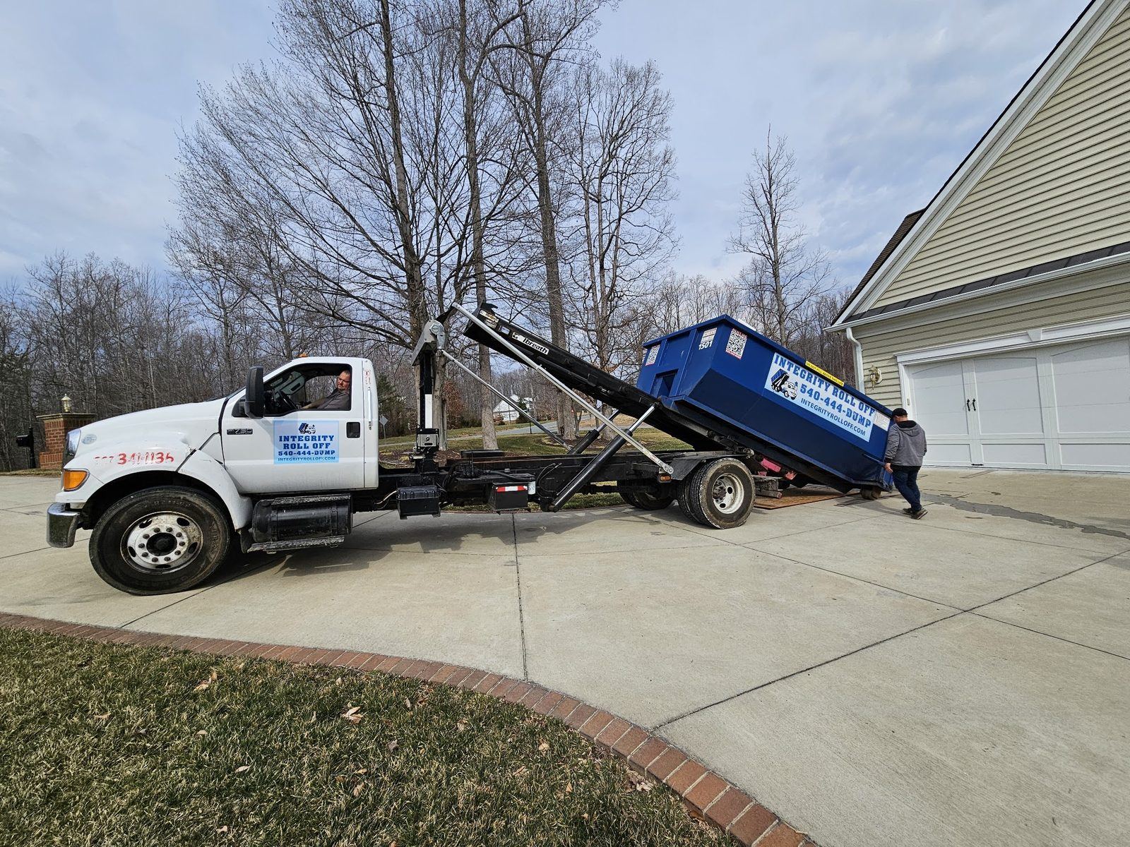 Integrity Roll Off truck delivering a blue roll off dumpster in a customer’s driveway